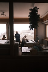 Several people are seated at tables in a cozy indoor setting with large windows providing a view of snow-capped mountains. A small decorative tree hangs from the ceiling, creating a festive atmosphere. The individuals appear to be engaged in conversation or enjoying a meal.