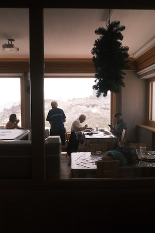 Several people are seated at tables in a cozy indoor setting with large windows providing a view of snow-capped mountains. A small decorative tree hangs from the ceiling, creating a festive atmosphere. The individuals appear to be engaged in conversation or enjoying a meal.