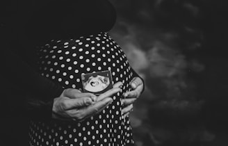 A black and white photograph of a pregnant person holding an ultrasound image in front of a polka dot dress. Hands gently cradle the belly, conveying a sense of care and anticipation.