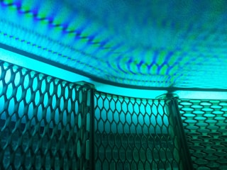 Intricate patterns of light and shadow cast on a textured surface, creating a rippling effect. A metallic mesh structure is positioned at an angle in the foreground, its hexagonal openings allowing light to filter through, resulting in a dynamic interplay of colors and forms.