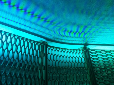 Intricate patterns of light and shadow cast on a textured surface, creating a rippling effect. A metallic mesh structure is positioned at an angle in the foreground, its hexagonal openings allowing light to filter through, resulting in a dynamic interplay of colors and forms.