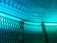 Intricate patterns of light and shadow cast on a textured surface, creating a rippling effect. A metallic mesh structure is positioned at an angle in the foreground, its hexagonal openings allowing light to filter through, resulting in a dynamic interplay of colors and forms.