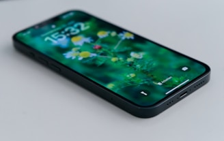 Smartphone placed on a flat surface with a close-up view of the screen displaying a vibrant floral wallpaper featuring daisies. The device is black, and the time displayed is 15:32. The image has a shallow depth of field, focusing mainly on the screen.