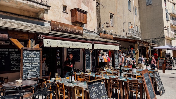 An outdoor restaurant setting located in what appears to be a quaint European town. The scene includes several wooden tables and chairs arranged in front of a restaurant with rustic signage. People are walking by, and menus are displayed on chalkboards. The building facade is aged, adding to the charming atmosphere.