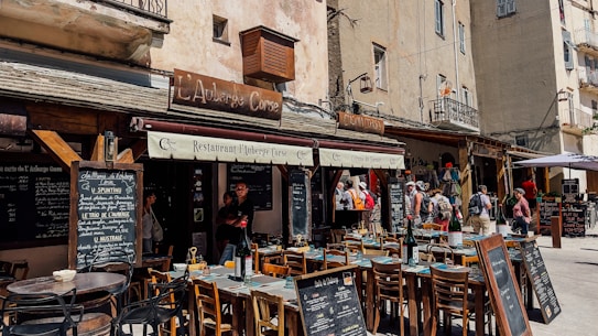 An outdoor restaurant setting located in what appears to be a quaint European town. The scene includes several wooden tables and chairs arranged in front of a restaurant with rustic signage. People are walking by, and menus are displayed on chalkboards. The building facade is aged, adding to the charming atmosphere.