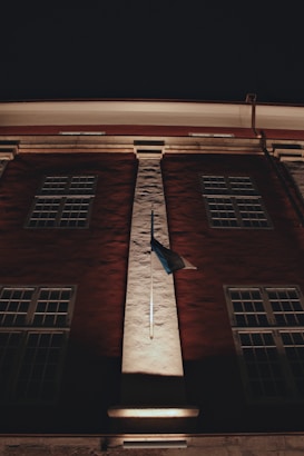 A tall building facade with multiple windows is illuminated at night. A flag is mounted on a central column, casting shadows that enhance its texture. The lighting highlights architectural details against a dark sky.