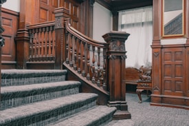 A wooden staircase with intricately carved railing and dark wood panels on the walls. The stairs are carpeted in a patterned gray material. A decorative bench with carved wooden features is seen in the background, near a curtained window. A large mirror with a gold frame is mounted on the wall to the right of the staircase.