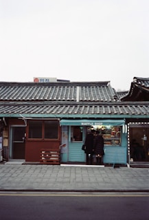 A quaint waffle shop with a traditional tiled roof set against an overcast sky. Two people are standing in front of the shop, possibly waiting for their order. The shop has a light blue facade and a matching bench outside.