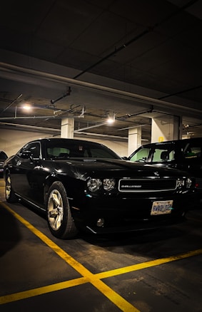 A black Dodge Challenger is parked in a dimly lit underground parking garage. The car's exterior is shiny and well-maintained, with visible chrome wheels. Overhead fluorescent lighting casts reflections on the car's surface. The garage has concrete pillars and a low ceiling with exposed pipes.
