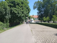 A pathway lined with lush green trees on the left, leading towards a building with a red-tiled roof in the background. Two people are walking down the path, with a cobblestone area to the right.