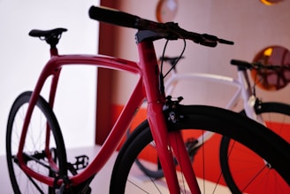 A close-up view of a sleek, modern bicycle with a red frame and black wheels, displayed in a well-lit showroom. In the background, another bicycle with a white frame is partially visible. The scene is set against a white and red wall with decorative circular elements.