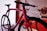 A close-up view of a sleek, modern bicycle with a red frame and black wheels, displayed in a well-lit showroom. In the background, another bicycle with a white frame is partially visible. The scene is set against a white and red wall with decorative circular elements.