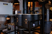 The image features an industrial setting with metal barrels arranged on a black rack. The barrels display a logo with a star and letter R. Surrounding equipment includes control panels and industrial machinery components.