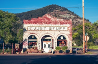 A quaint old-fashioned store displays goods outside, set against the backdrop of a mountain and greenery. The building has a rustic, brick facade with vintage signage. An American flag is displayed on the front porch.