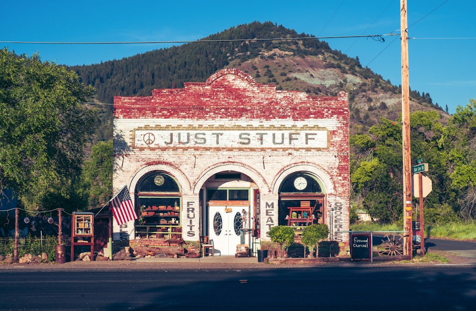 A quaint old-fashioned store displays goods outside, set against the backdrop of a mountain and greenery. The building has a rustic, brick facade with vintage signage. An American flag is displayed on the front porch.