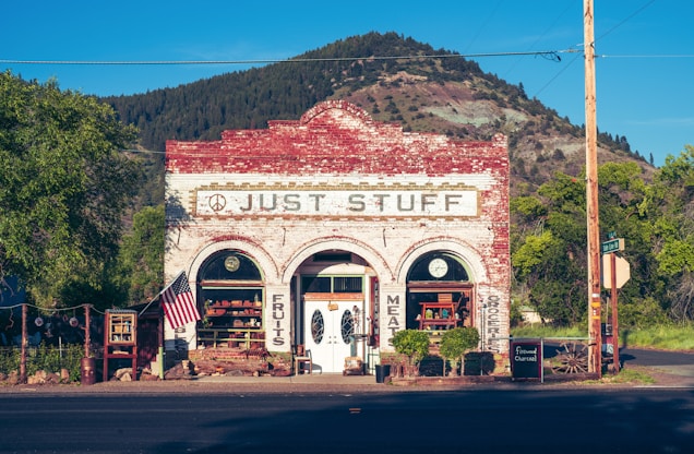 A quaint old-fashioned store displays goods outside, set against the backdrop of a mountain and greenery. The building has a rustic, brick facade with vintage signage. An American flag is displayed on the front porch.