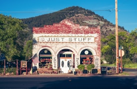 A quaint old-fashioned store displays goods outside, set against the backdrop of a mountain and greenery. The building has a rustic, brick facade with vintage signage. An American flag is displayed on the front porch.