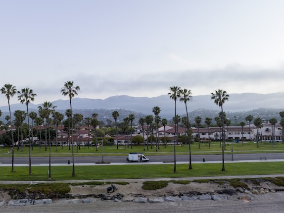 A coastal scene featuring a well-maintained park with palm trees. In the background, there are residential buildings with red roofs and mountains. A white van is driving on a road that separates the park from the residential area. The sky is partly cloudy, and the area near the ocean is rocky and sandy.