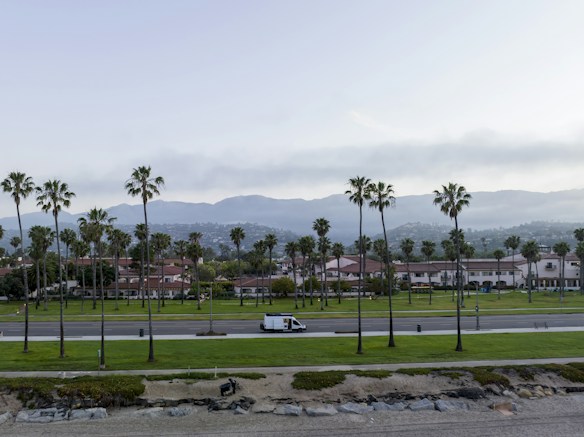 A coastal scene featuring a well-maintained park with palm trees. In the background, there are residential buildings with red roofs and mountains. A white van is driving on a road that separates the park from the residential area. The sky is partly cloudy, and the area near the ocean is rocky and sandy.
