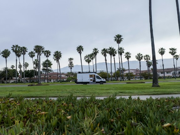 A white van is parked on a grassy area surrounded by tall palm trees, with a backdrop of mountains under an overcast sky. Residential houses with red-tiled roofs are visible in the distance, and the scene has a calm and serene atmosphere.