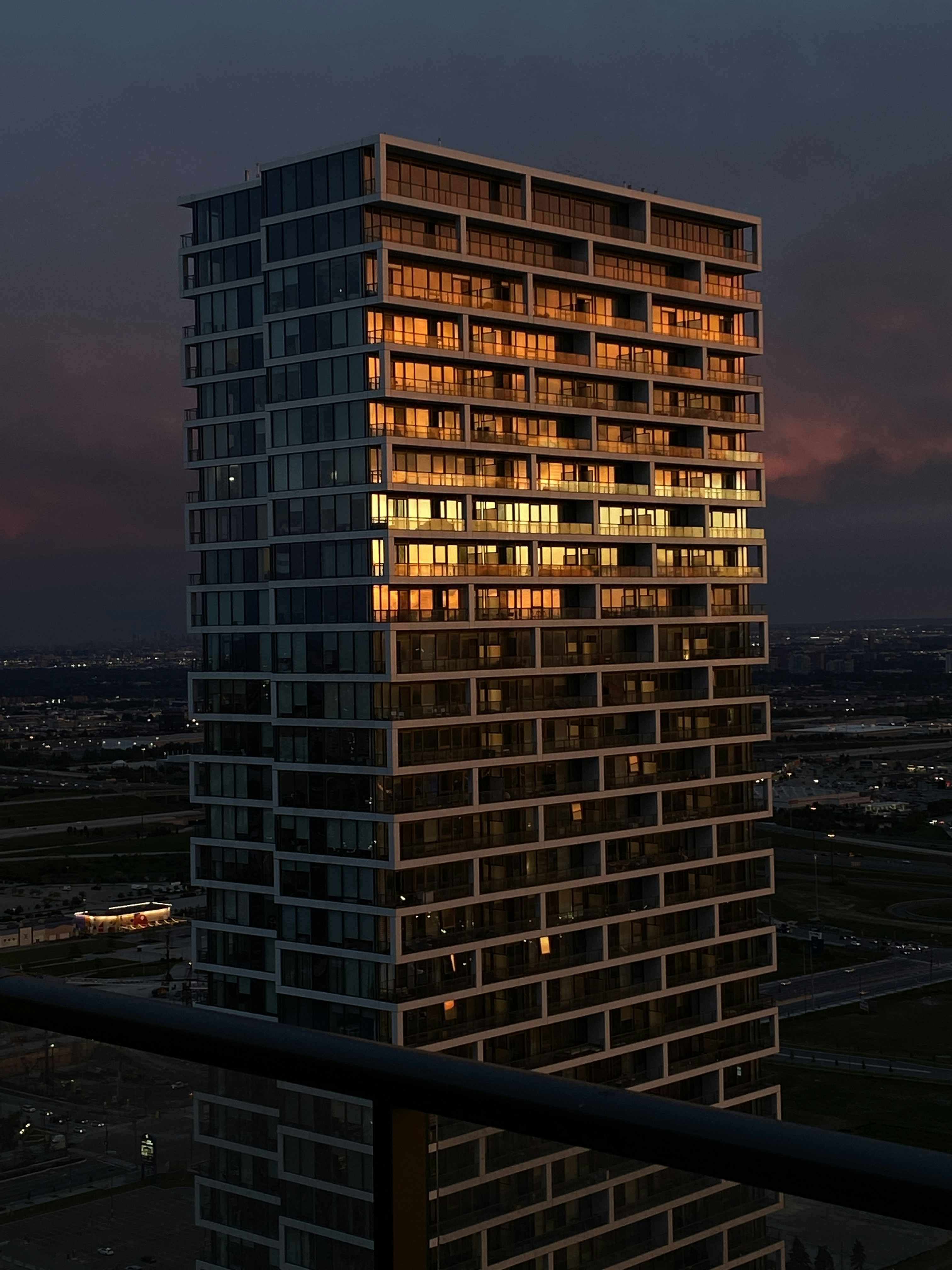 Modern glass office tower at dusk with glowing windows, dark sky, dramatic urban lighting, deep shadows on facade