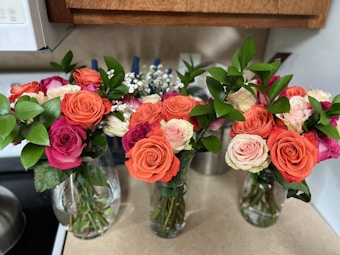 Three glass vases are filled with vibrant bouquets of roses in various colors, including orange, pink, and cream. Fresh green leaves and small white filler flowers complement the roses. The arrangement is set on a kitchen countertop with a neutral backsplash and wooden cabinets above.