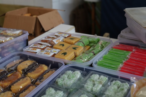Packages of assorted snacks are arranged in clear plastic containers on a table. The snacks vary in colors and shapes, including green, red, and yellow items wrapped in clear plastic. There is also a cardboard box in the background with more wrapped snacks and some unwrapped items visible.