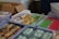 Packages of assorted snacks are arranged in clear plastic containers on a table. The snacks vary in colors and shapes, including green, red, and yellow items wrapped in clear plastic. There is also a cardboard box in the background with more wrapped snacks and some unwrapped items visible.