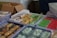 Packages of assorted snacks are arranged in clear plastic containers on a table. The snacks vary in colors and shapes, including green, red, and yellow items wrapped in clear plastic. There is also a cardboard box in the background with more wrapped snacks and some unwrapped items visible.