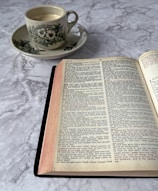 An open book on a marble surface with a cup of coffee in a floral-patterned saucer nearby. The pages of the book contain text in two columns, resembling a religious or historical document. The coffee cup appears to be filled with a light-colored beverage.