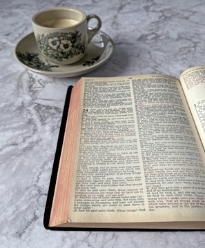 An open book on a marble surface with a cup of coffee in a floral-patterned saucer nearby. The pages of the book contain text in two columns, resembling a religious or historical document. The coffee cup appears to be filled with a light-colored beverage.
