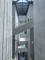 A modern architectural space with high ceilings and large windows allowing natural light to illuminate the interior. Several people are walking along a glass-railed walkway that crosses between two tall, concrete walls. The design is open and minimalistic, with an emphasis on vertical lines and shadows.