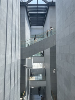 A modern architectural space with high ceilings and large windows allowing natural light to illuminate the interior. Several people are walking along a glass-railed walkway that crosses between two tall, concrete walls. The design is open and minimalistic, with an emphasis on vertical lines and shadows.