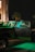 A truck is parked at night with its doors open, illuminated by green light. The truck is covered with various graphics and logos. A helmet can be seen on the driver's seat. The setting includes palm trees and the exterior of a building in the background.