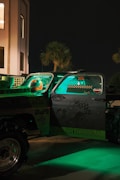 A truck is parked at night with its doors open, illuminated by green light. The truck is covered with various graphics and logos. A helmet can be seen on the driver's seat. The setting includes palm trees and the exterior of a building in the background.
