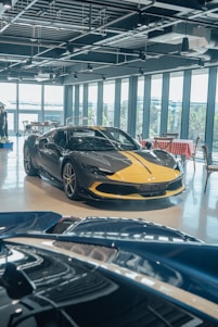 A luxury sports car with a sleek black and yellow design is displayed in a modern showroom with large windows. Indoor lighting creates reflections on the polished surfaces, and a light blue convertible car is partially visible in the foreground. The showroom has a contemporary ambiance with exposed ceiling fixtures and furniture with red and white checkered tablecloths.