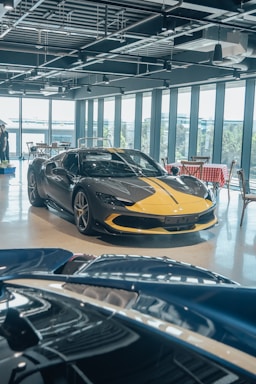 A luxury sports car with a sleek black and yellow design is displayed in a modern showroom with large windows. Indoor lighting creates reflections on the polished surfaces, and a light blue convertible car is partially visible in the foreground. The showroom has a contemporary ambiance with exposed ceiling fixtures and furniture with red and white checkered tablecloths.