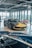 A luxury sports car with a sleek black and yellow design is displayed in a modern showroom with large windows. Indoor lighting creates reflections on the polished surfaces, and a light blue convertible car is partially visible in the foreground. The showroom has a contemporary ambiance with exposed ceiling fixtures and furniture with red and white checkered tablecloths.