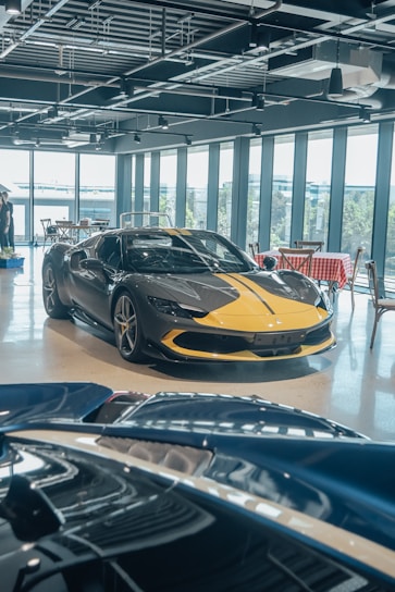 A luxury sports car with a sleek black and yellow design is displayed in a modern showroom with large windows. Indoor lighting creates reflections on the polished surfaces, and a light blue convertible car is partially visible in the foreground. The showroom has a contemporary ambiance with exposed ceiling fixtures and furniture with red and white checkered tablecloths.