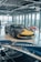 A luxury sports car with a sleek black and yellow design is displayed in a modern showroom with large windows. Indoor lighting creates reflections on the polished surfaces, and a light blue convertible car is partially visible in the foreground. The showroom has a contemporary ambiance with exposed ceiling fixtures and furniture with red and white checkered tablecloths.