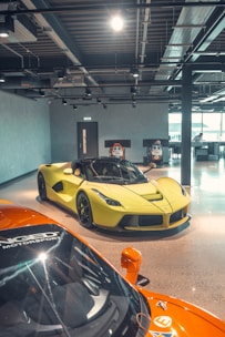 A sleek, yellow sports car is prominently displayed in a modern indoor showroom with a polished concrete floor. In the foreground, the side of a bright orange sports car is partially visible. The space features industrial-style lighting and visible ductwork on the ceiling, with large windows allowing natural light to fill the area. Racing simulators are positioned against the wall in the background.