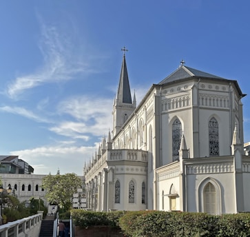 Gothic-style white church with a tall steeple reaching towards the clear blue sky. The church features arched stained-glass windows and intricate detailing. Surrounding the church are lush green shrubs, signage, and pathways, with one pathway having steps leading up. In the background, modern buildings contrast with the historic architecture of the church.