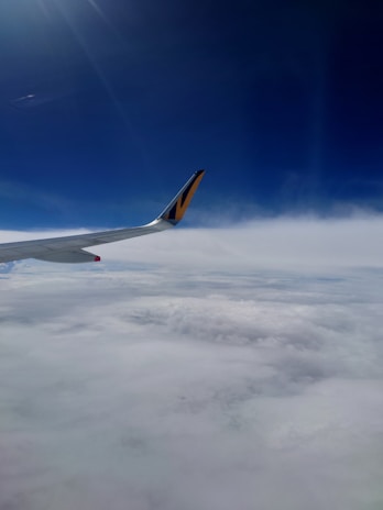 An airplane wing with a distinctive yellow and black design extends across the view, set against a backdrop of blue sky and thick white clouds below.