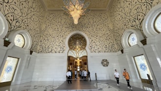 An opulent interior space features intricate floral patterns on grand, arched walls and a soaring ceiling. Large, ornate chandeliers hang from above, illuminating the space. The floor and walls are made of glossy white materials, enhancing the luxurious feel. Visitors are visible within the room, adding a sense of scale to the impressive architecture.