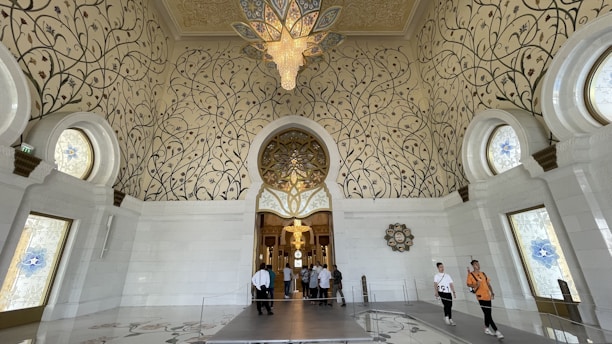 An opulent interior space features intricate floral patterns on grand, arched walls and a soaring ceiling. Large, ornate chandeliers hang from above, illuminating the space. The floor and walls are made of glossy white materials, enhancing the luxurious feel. Visitors are visible within the room, adding a sense of scale to the impressive architecture.