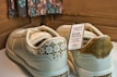 A pair of cream-colored sneakers with decorative patterns on the heel and a tag attached indicating the Batik Series. The tag includes text about the brand and style, highlighting collaboration and availability. The background features a patterned fabric and wooden panels.