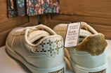 A pair of cream-colored sneakers with decorative patterns on the heel and a tag attached indicating the Batik Series. The tag includes text about the brand and style, highlighting collaboration and availability. The background features a patterned fabric and wooden panels.
