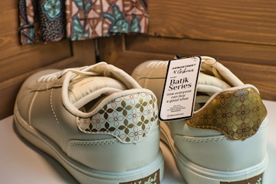 A pair of cream-colored sneakers with decorative patterns on the heel and a tag attached indicating the Batik Series. The tag includes text about the brand and style, highlighting collaboration and availability. The background features a patterned fabric and wooden panels.