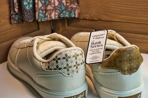 A pair of cream-colored sneakers with decorative patterns on the heel and a tag attached indicating the Batik Series. The tag includes text about the brand and style, highlighting collaboration and availability. The background features a patterned fabric and wooden panels.
