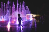 A silhouette of a child running through an illuminated fountain at night. The water jets create a vibrant and colorful display with shades of purple, blue, and pink. In the background, there are people observing the scene, and a well-lit building is visible.